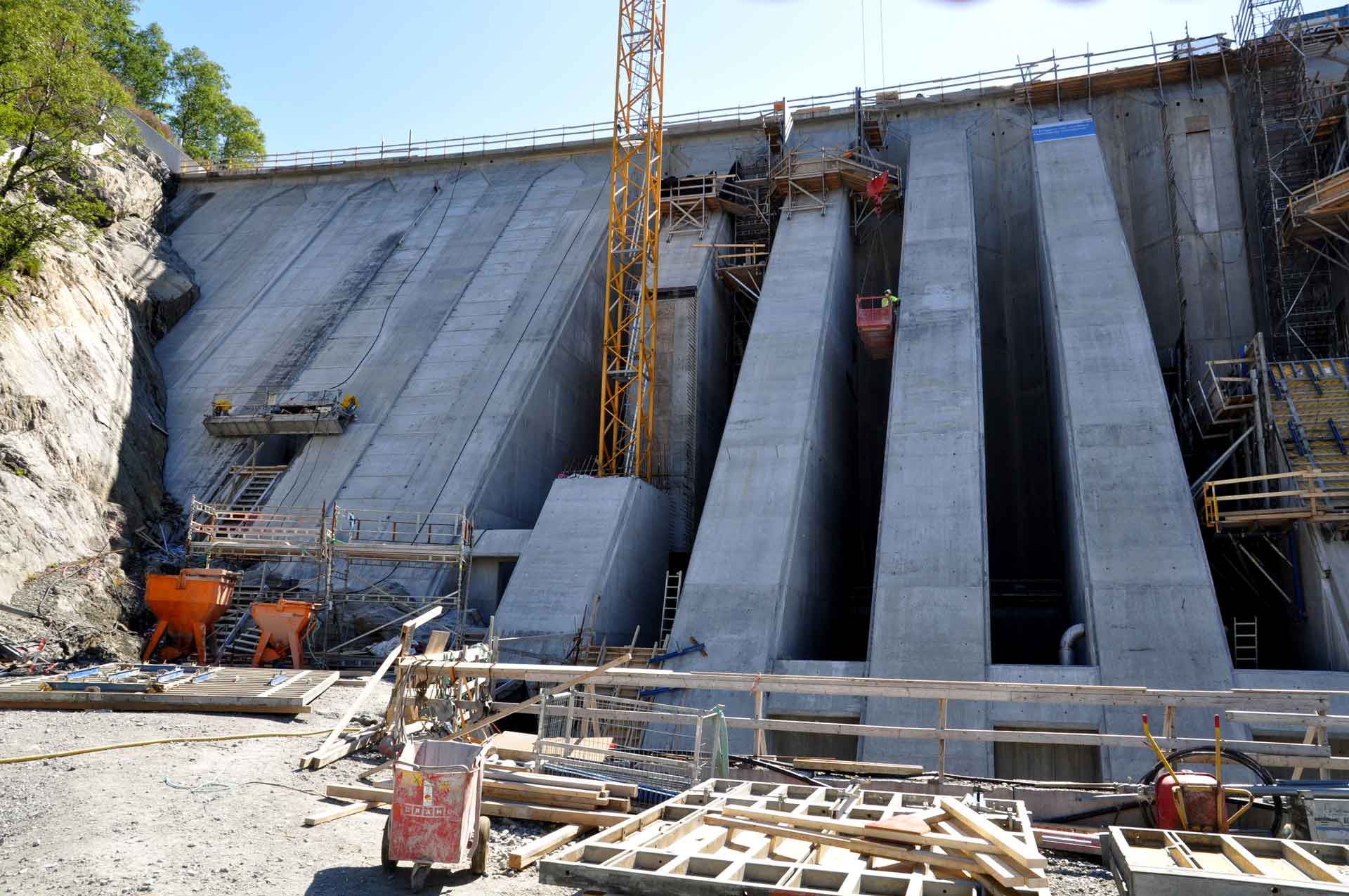 A large concrete dam under construction features several tall, vertical concrete walls with scaffolding and construction machinery, including cranes and an orange excavator at the foot of a rocky slope.