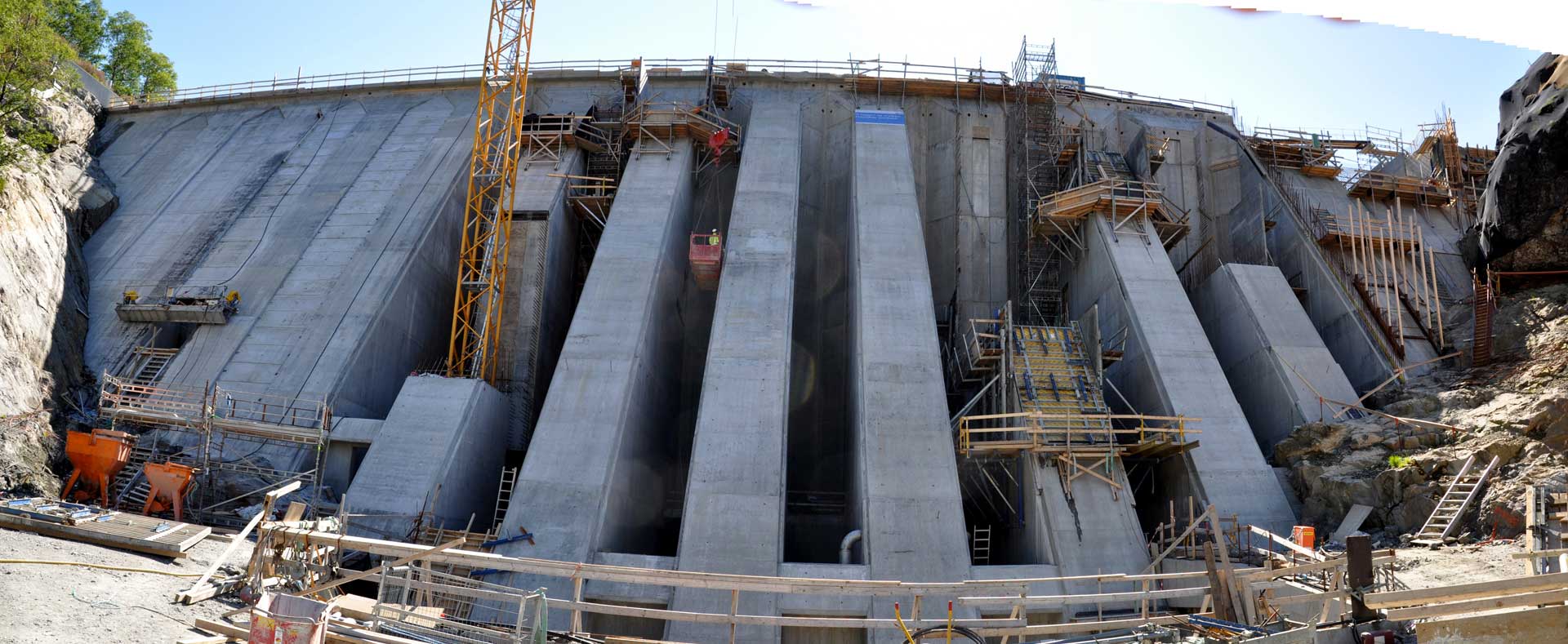 A large concrete dam under construction featuring numerous support structures and construction machinery, set against a backdrop of rocky terrain beneath a clear sky.