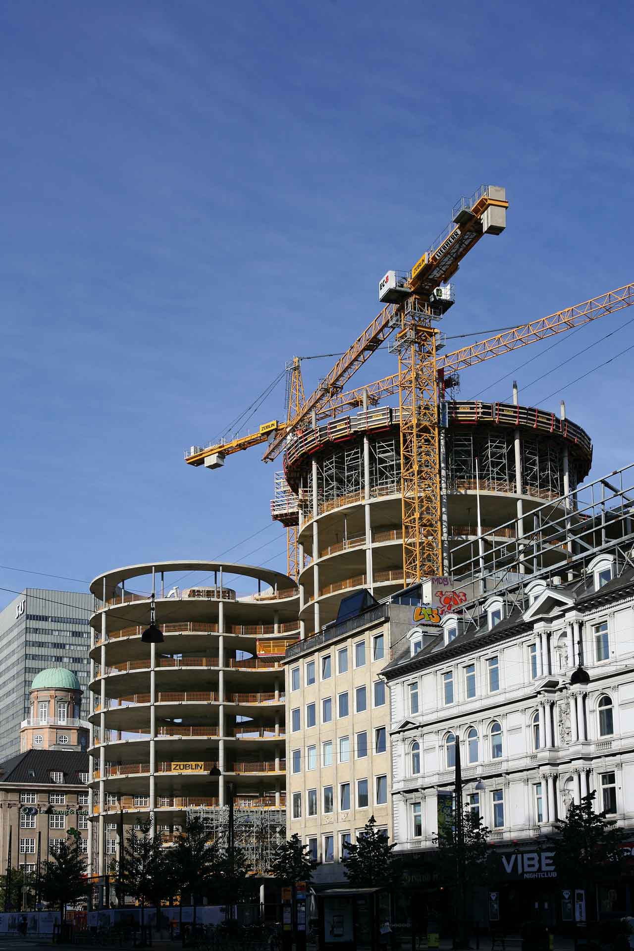 A construction site with a large yellow crane towering over a partially constructed multi-story building. In the foreground, there are traditional buildings with classic architectural designs, indicating that the construction is taking place in an urban area blending new developments with old city structures.