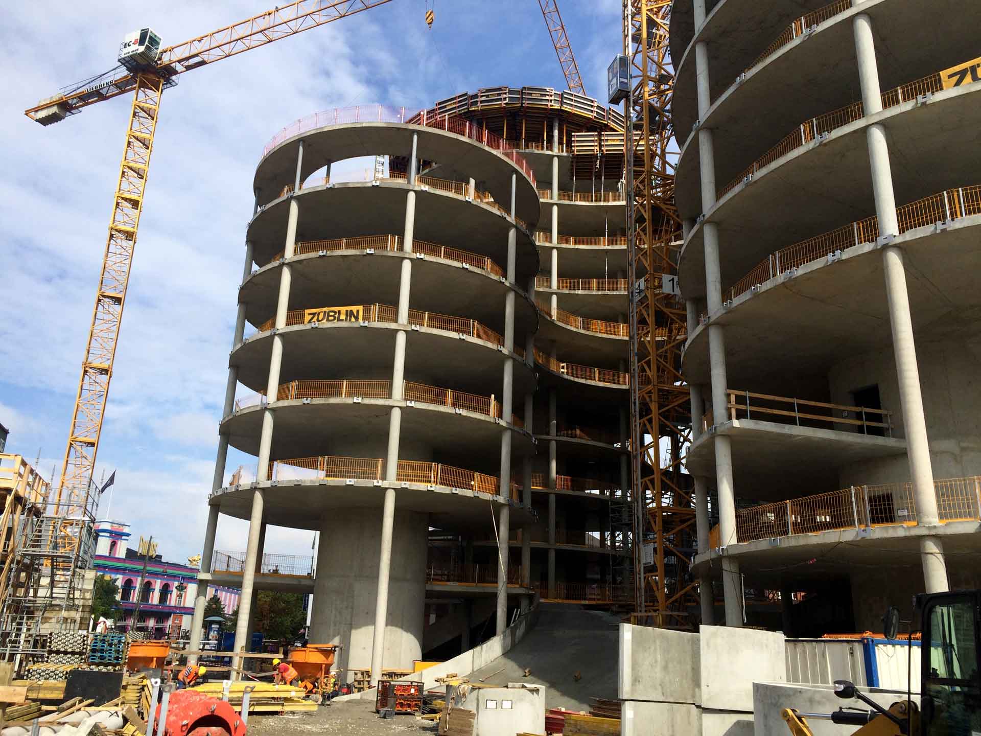 A construction site with a multi-story building under construction. The building has a curved design with multiple floors visible.