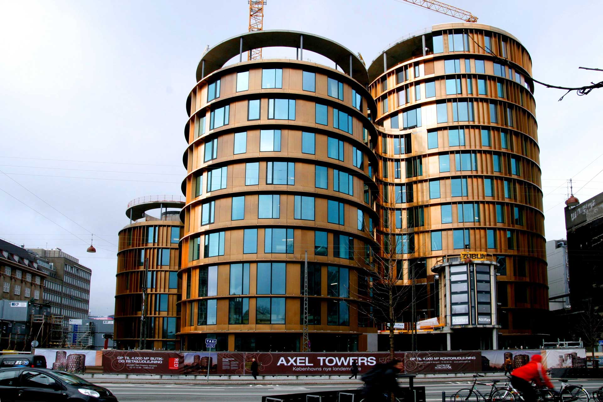 A modern building under construction with a distinctive architectural design featuring curved shapes and a facade with gold and brown hues. The building has multiple floors, large windows, and two visible cranes on top, indicating ongoing work.