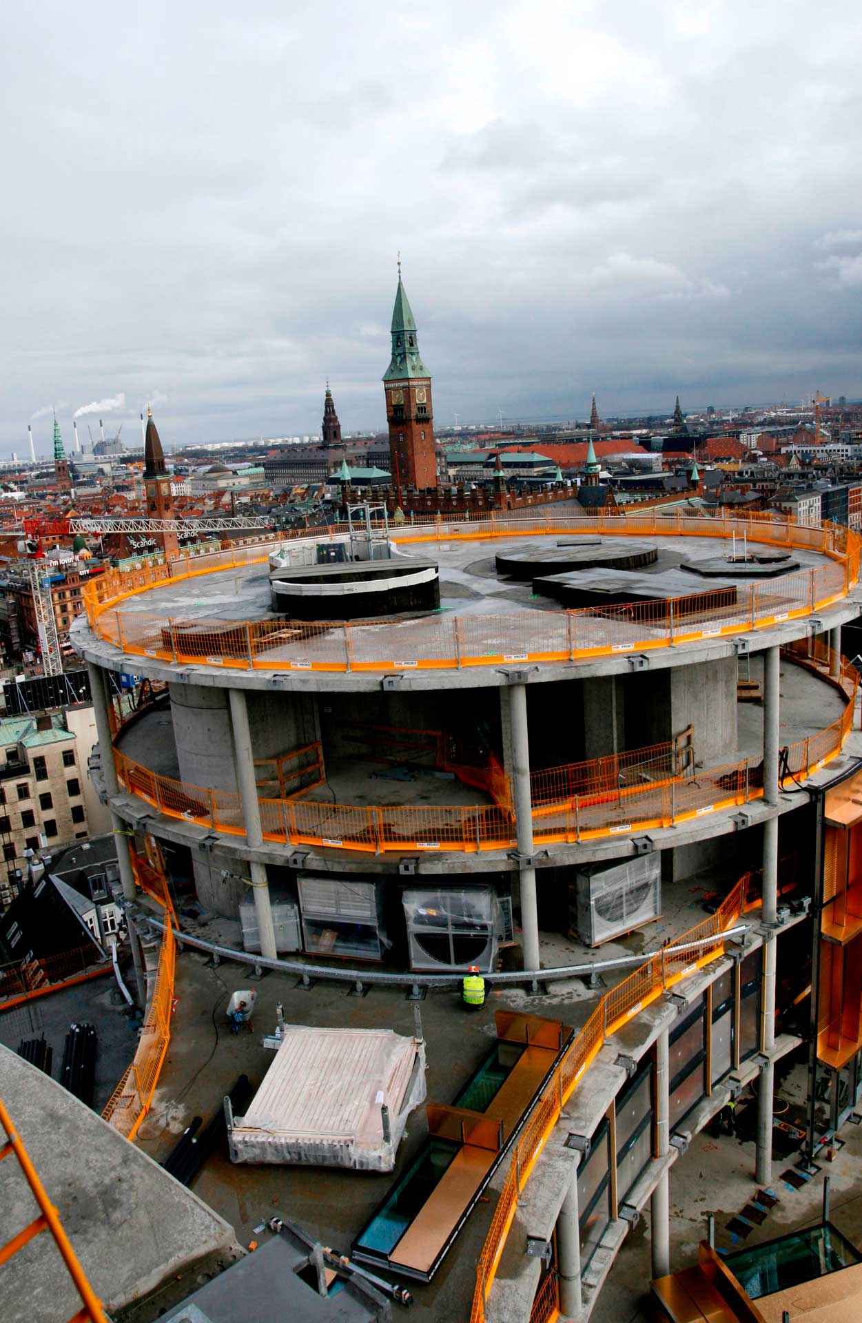 A large, multi-level circular construction site with orange safety barriers. The construction is surrounded by an urban environment with buildings in the background, including a notable tower with a green roof and spire.