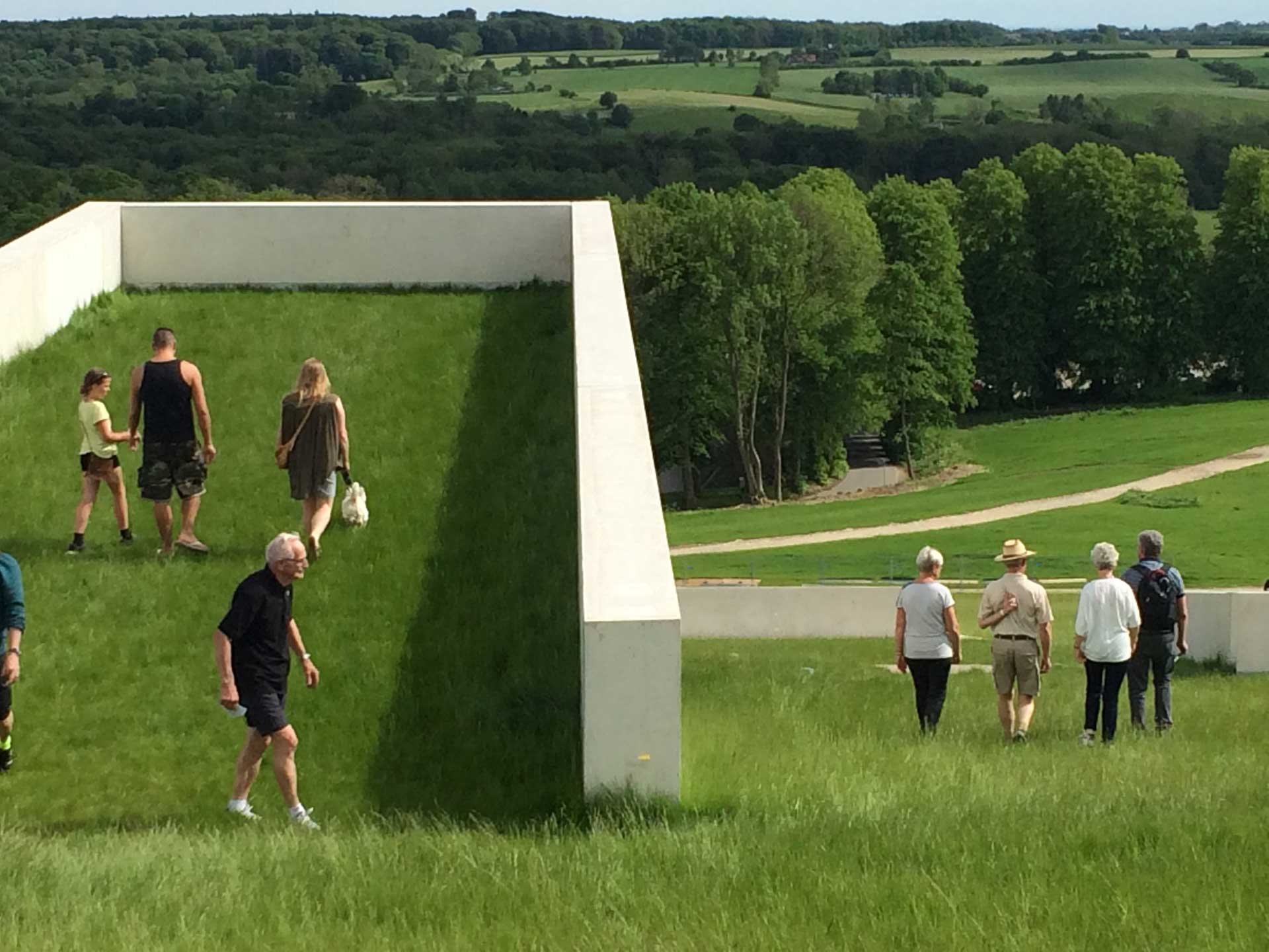 A large white cubic sculpture in a lush field with people walking around and inside it, under a clear blue sky.