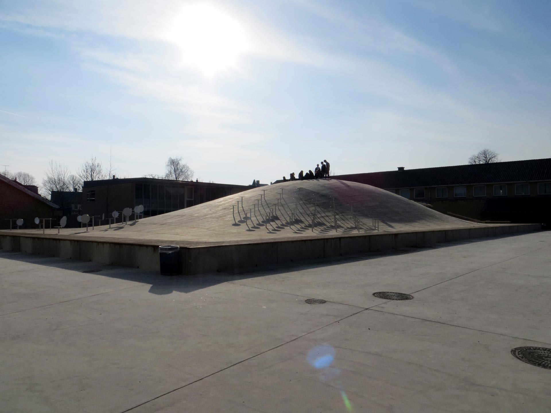 A concrete dome with a smooth surface rises in the center of a paved square, surrounded by low buildings under a clear sky. Several individuals stand atop the dome, casting shadows onto the curved surface.