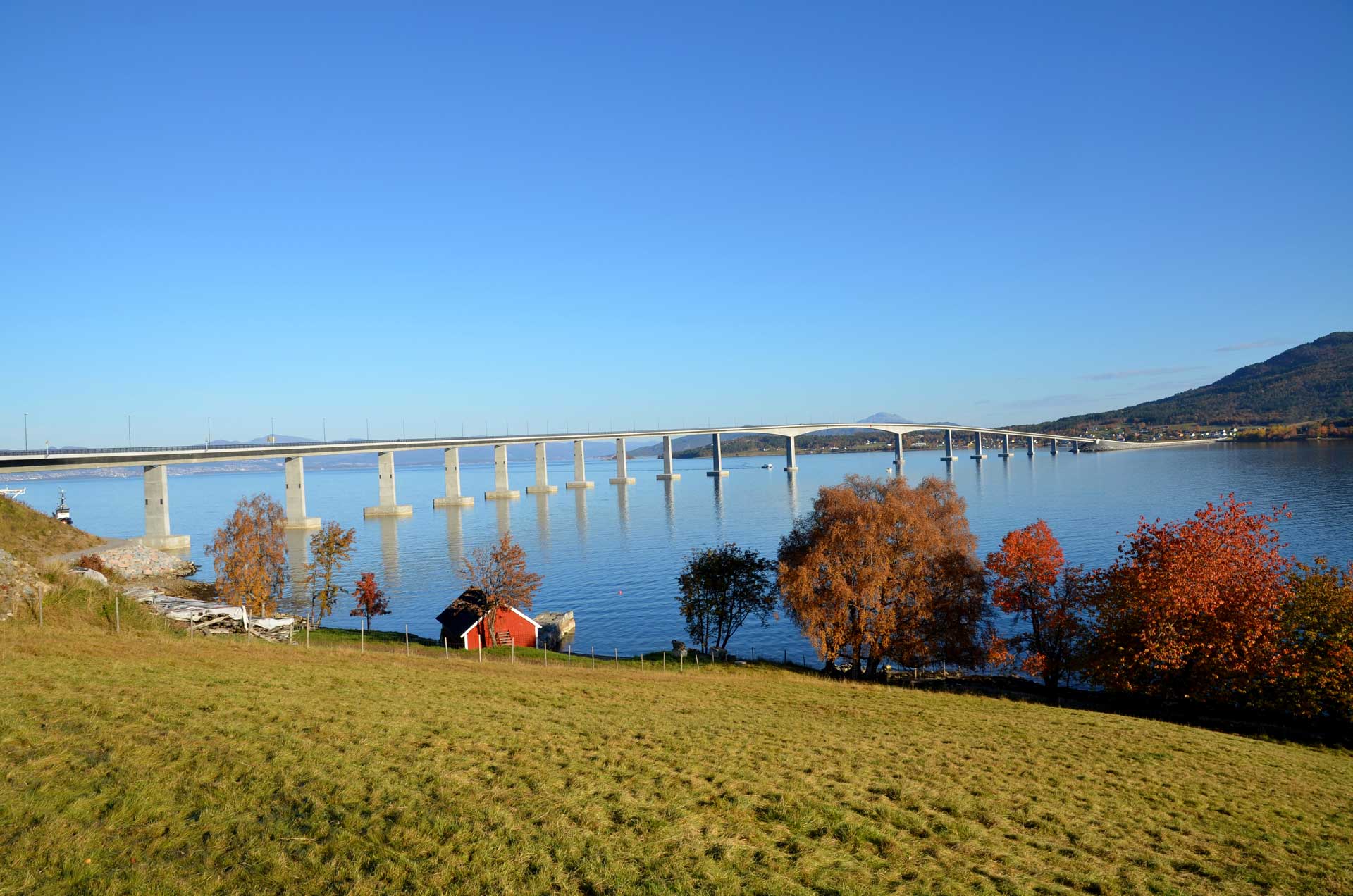 A long bridge spans a large body of water. In the foreground, there is a grassy area with trees in autumnal colors and a small red house with a black roof near the shore.