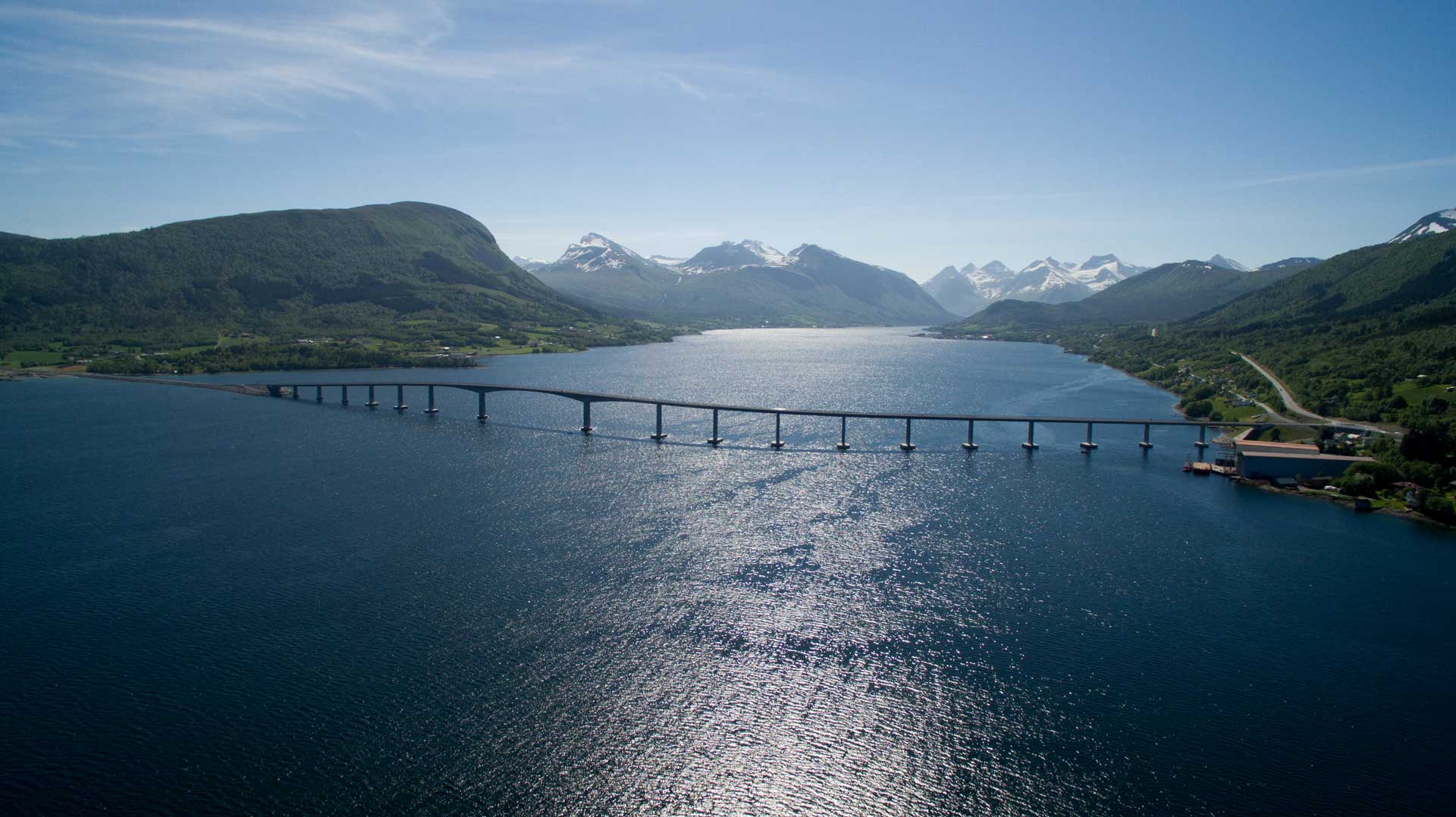 A bridge spans a wide body of water, flanked by green shores and mountains in the background. The shadow of the bridge is clearly cast upon the surface of the water.