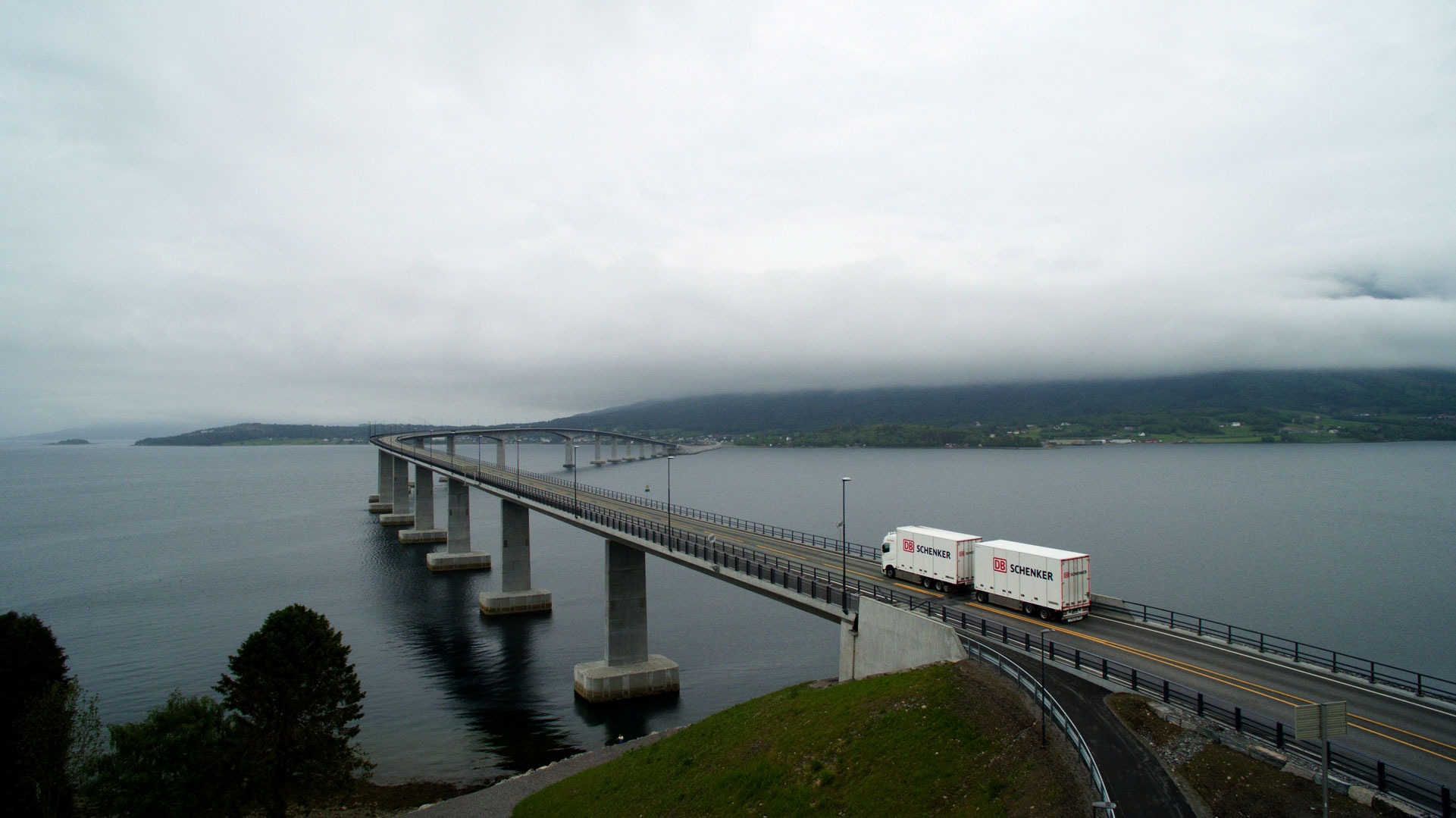 A long bridge extends over a body of water with a truck driving on it. The bridge connects two land masses, and the weather is overcast with low-lying clouds or fog.