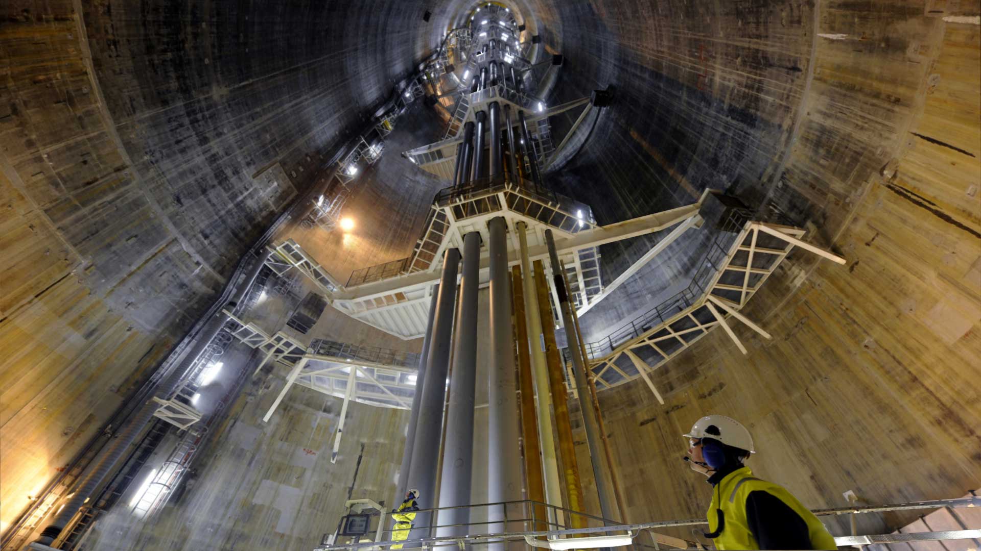 The interior of the Troll A oil platform, a large cylindrical structure, with multiple metal beams and scaffolding. A worker wearing a hard hat and reflective vest is visible in the foreground, indicating an industrial or construction setting.