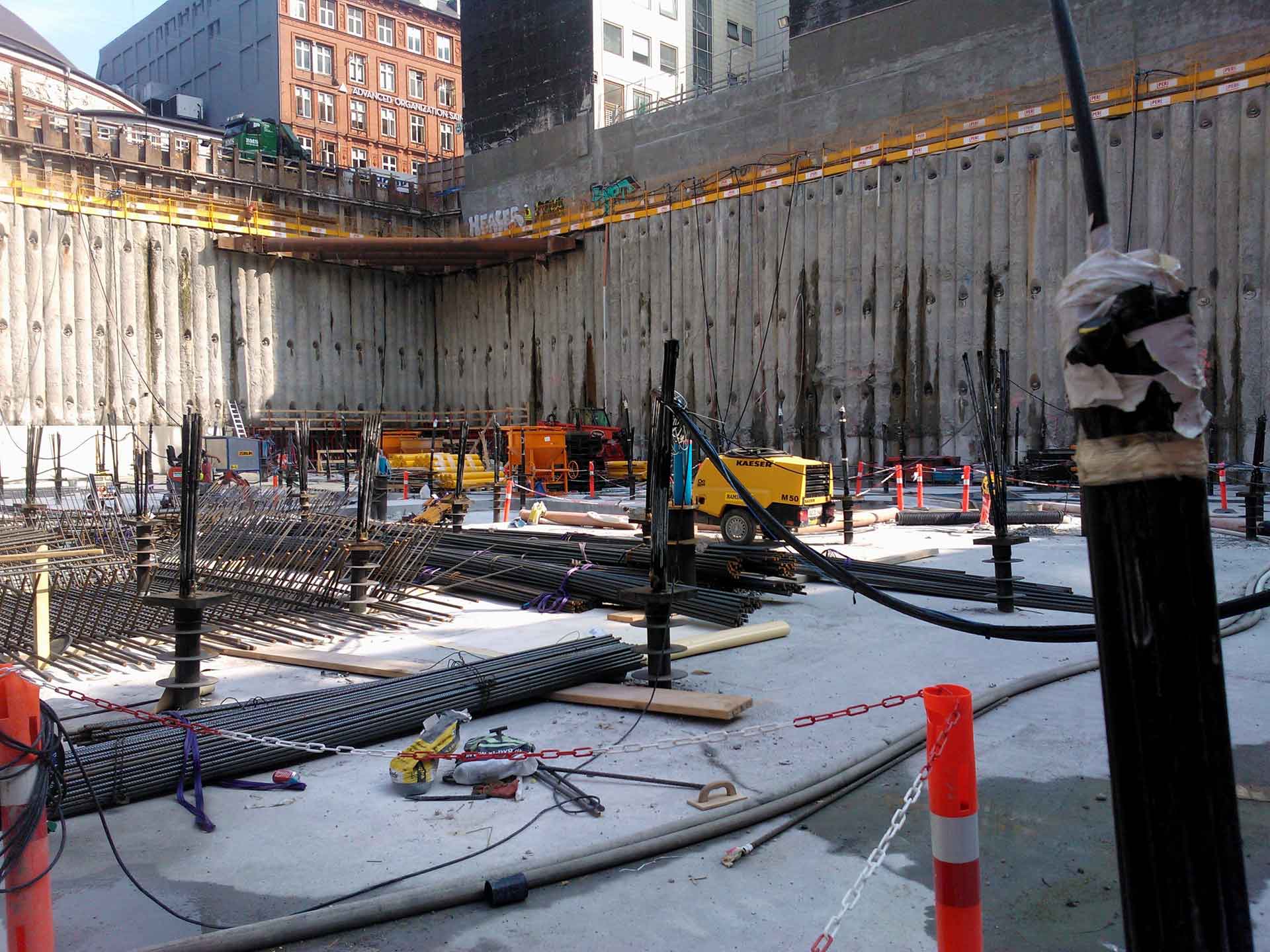 A construction site with a large excavation pit. There are reinforced steel bars laid out on the ground, heavy machinery such as an excavator and a crane, and various construction materials scattered around.