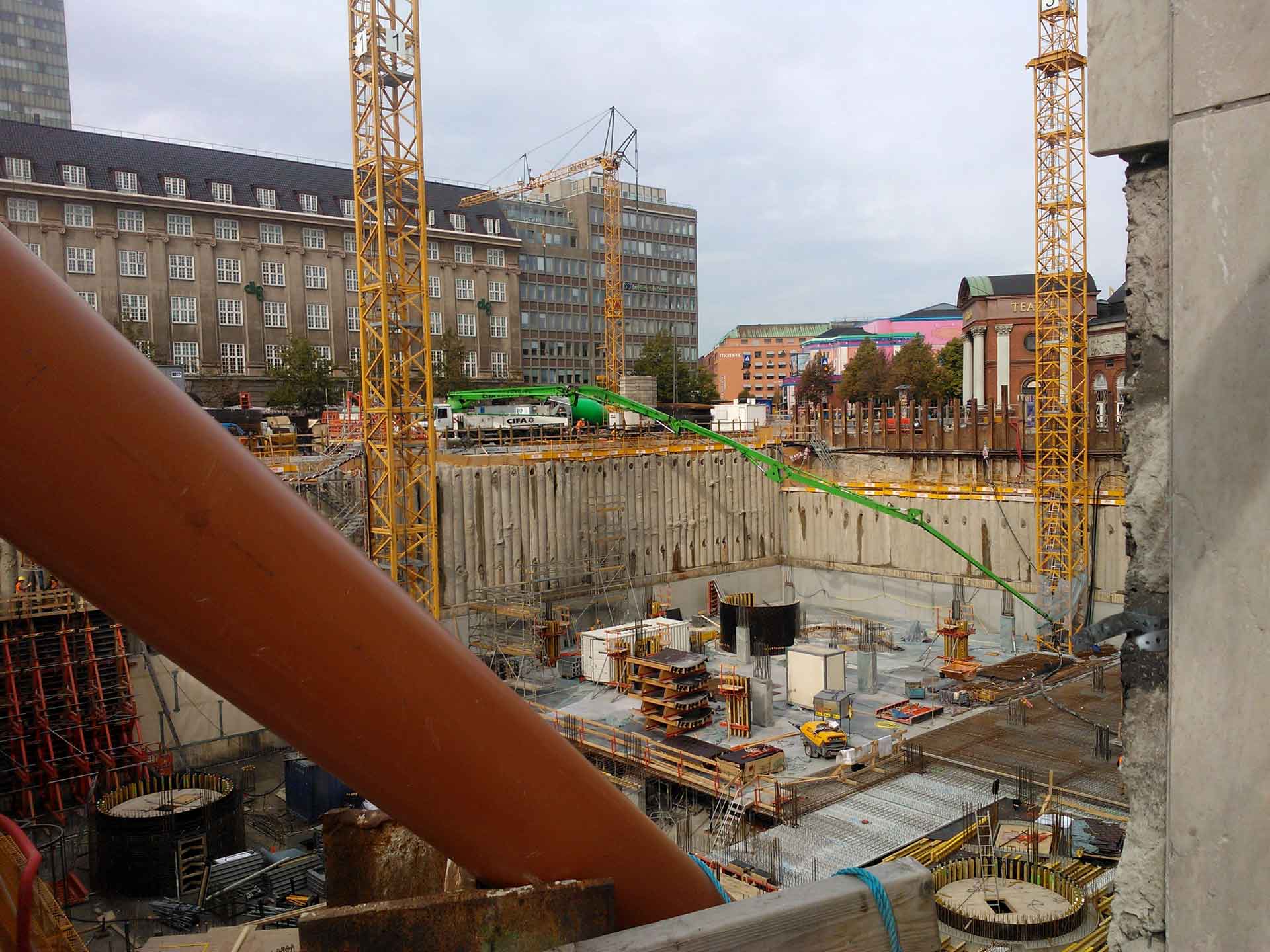 A construction site with multiple cranes, a deep excavation pit with retaining walls, and various construction materials scattered around the site. Buildings are visible in the background, indicating that this construction is taking place in an urban area.