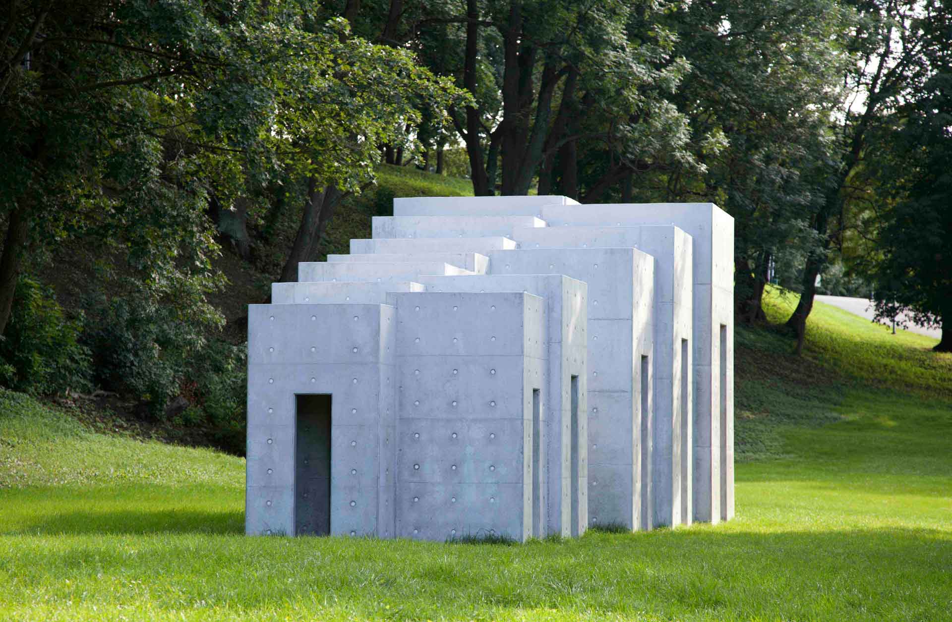 A white concrete sculpture with multiple rectangular sections arranged in a stacked, overlapping pattern stands on a grassy slope in a park. Trees are visible in the background.