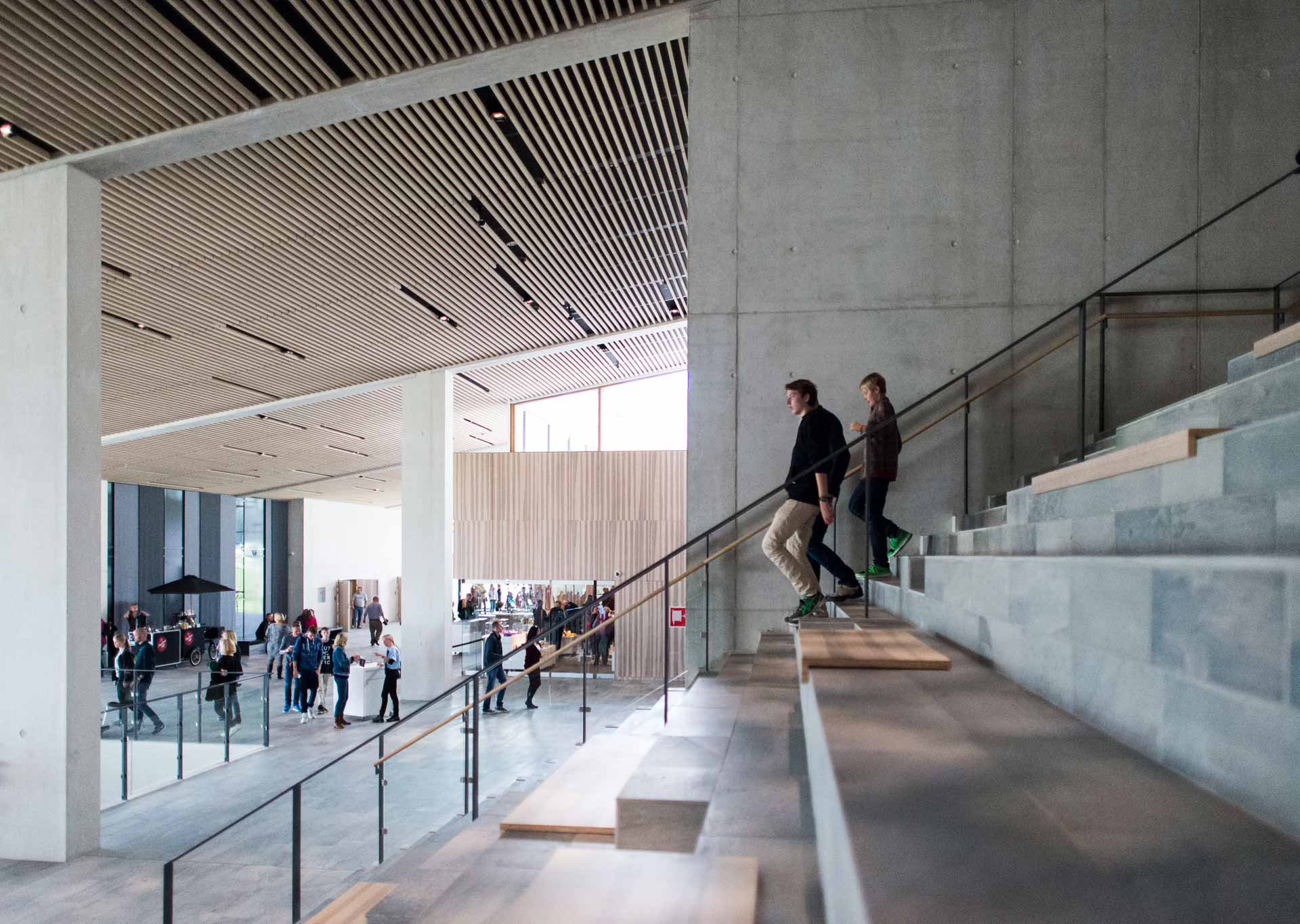 An interior space showcases modern architecture with a staircase that has glass balustrades and concrete steps. People are ascending the staircase; below is an expansive area with more people seated at tables.