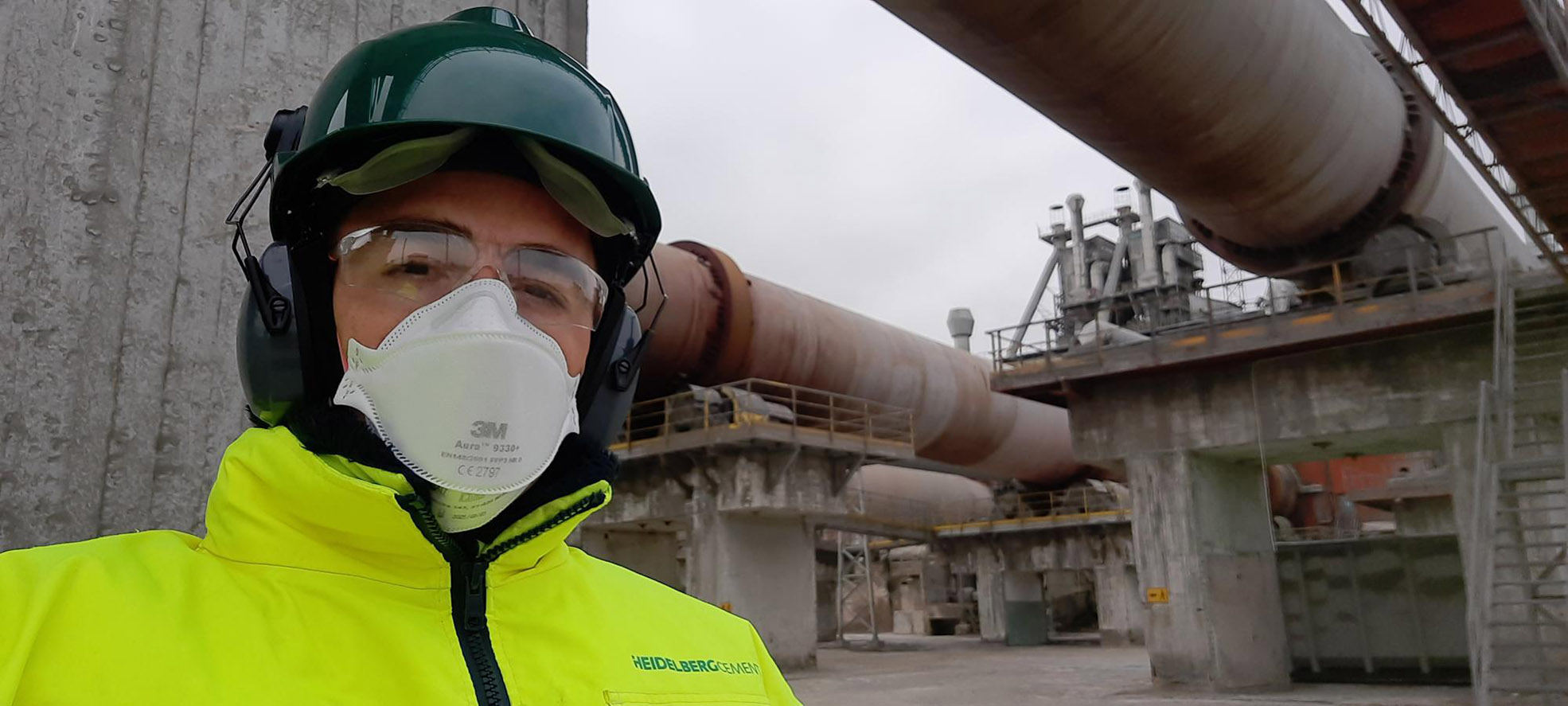 Man wearing face mask and protective gear in front of large tubes on a cement plant