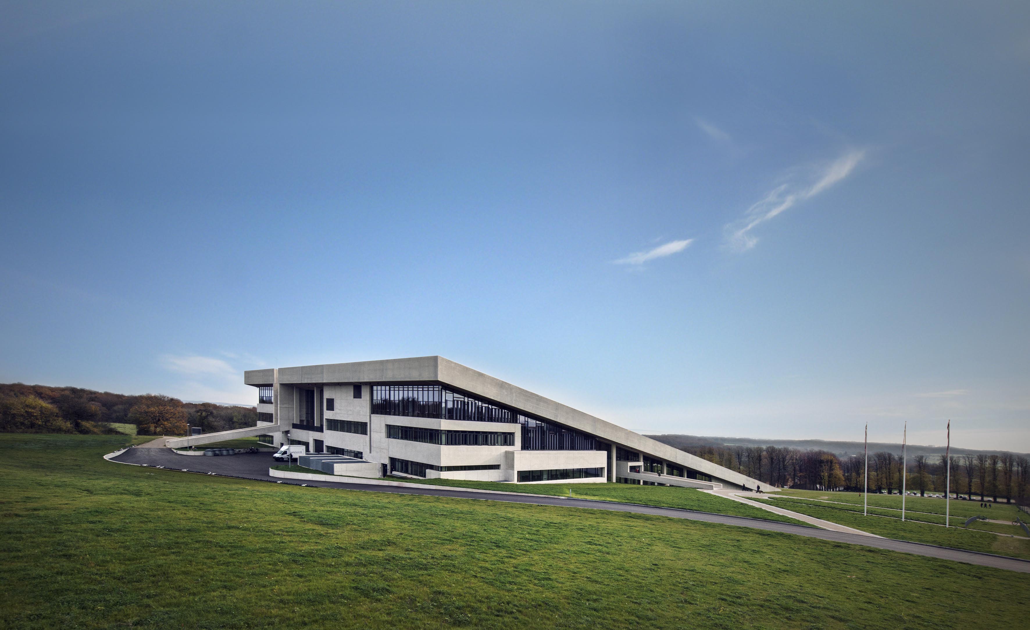 A modern building with a unique architectural design, featuring a prominent sloped roof that extends down to the ground level on one side. The building is situated on a grassy hill under a clear blue sky with some wisps of clouds.