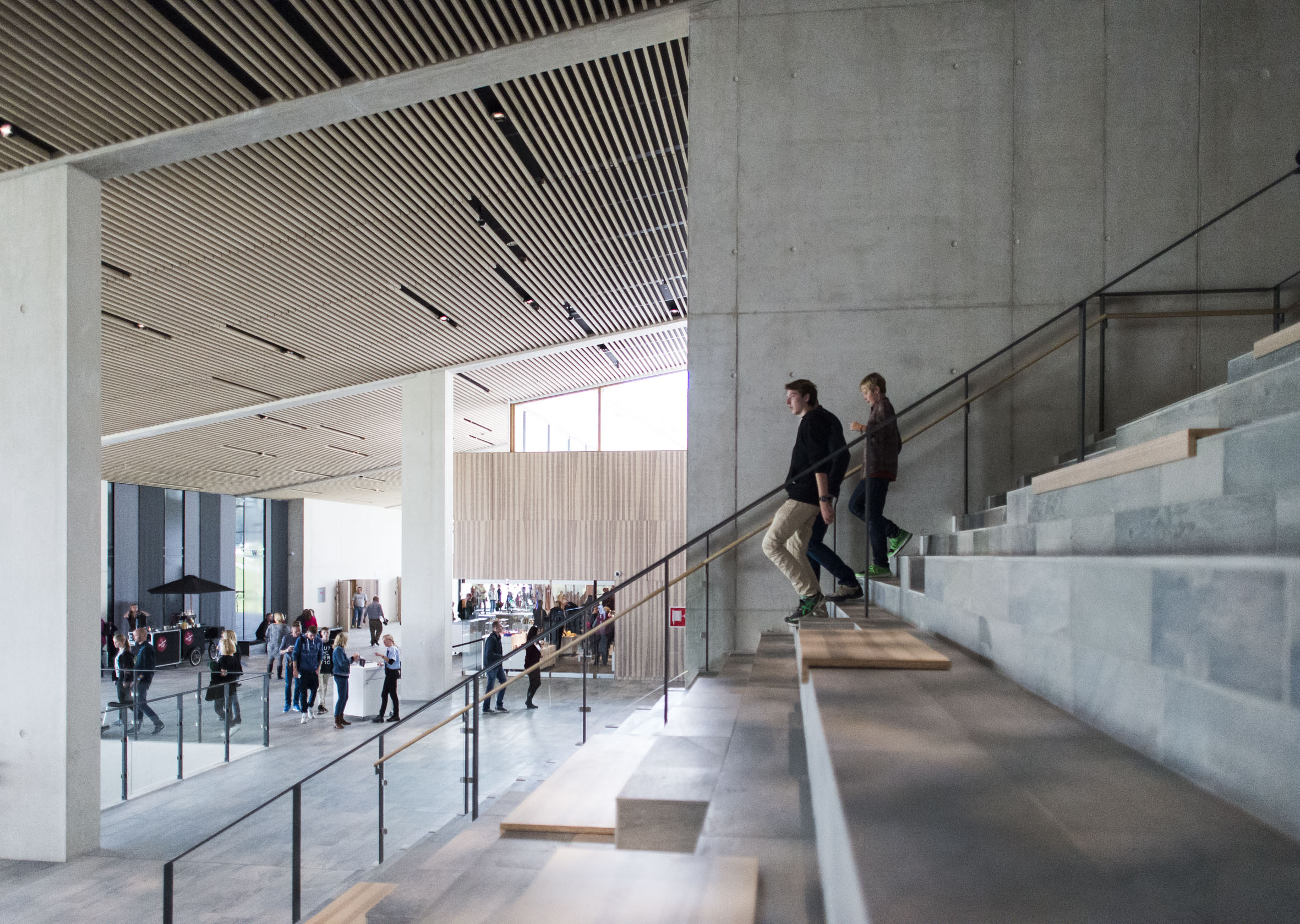 An interior space showcases modern architecture with a staircase that has glass balustrades and concrete steps. People are ascending the staircase; below is an expansive area with more people seated at tables.