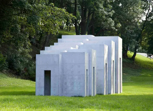 A white concrete sculpture with multiple rectangular sections arranged in a stacked, overlapping pattern stands on a grassy slope in a park. Trees are visible in the background.